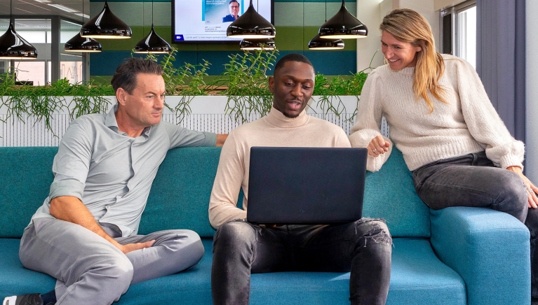 Drie medewerkers van GBLT overleggen met elkaar op een bank op kantoor. Ze kijken naar een laptop